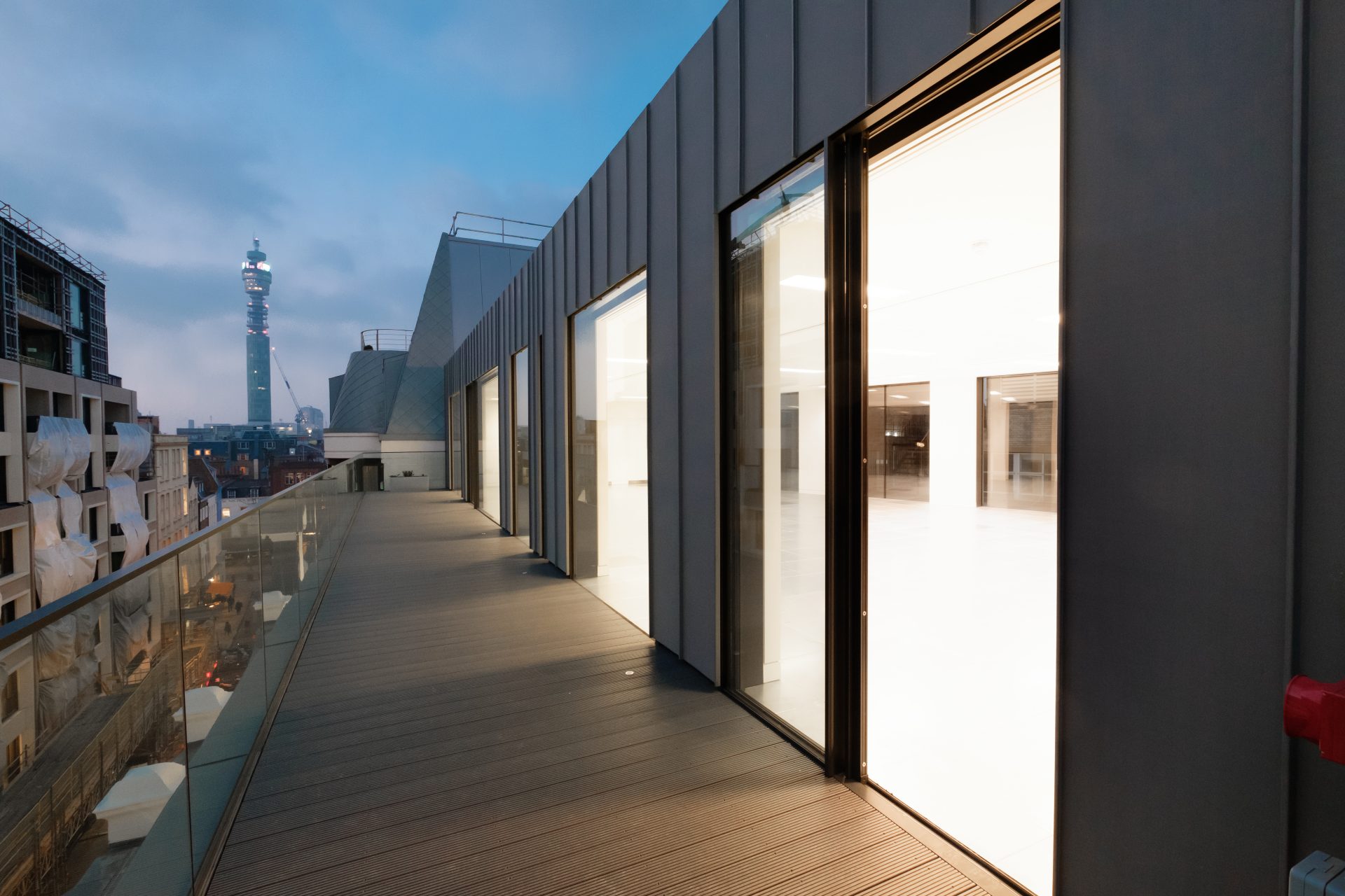 modern architectural entrance featuring a glass walkway with a glass balustrade leading to a building with dark grey walls and sliding doors.